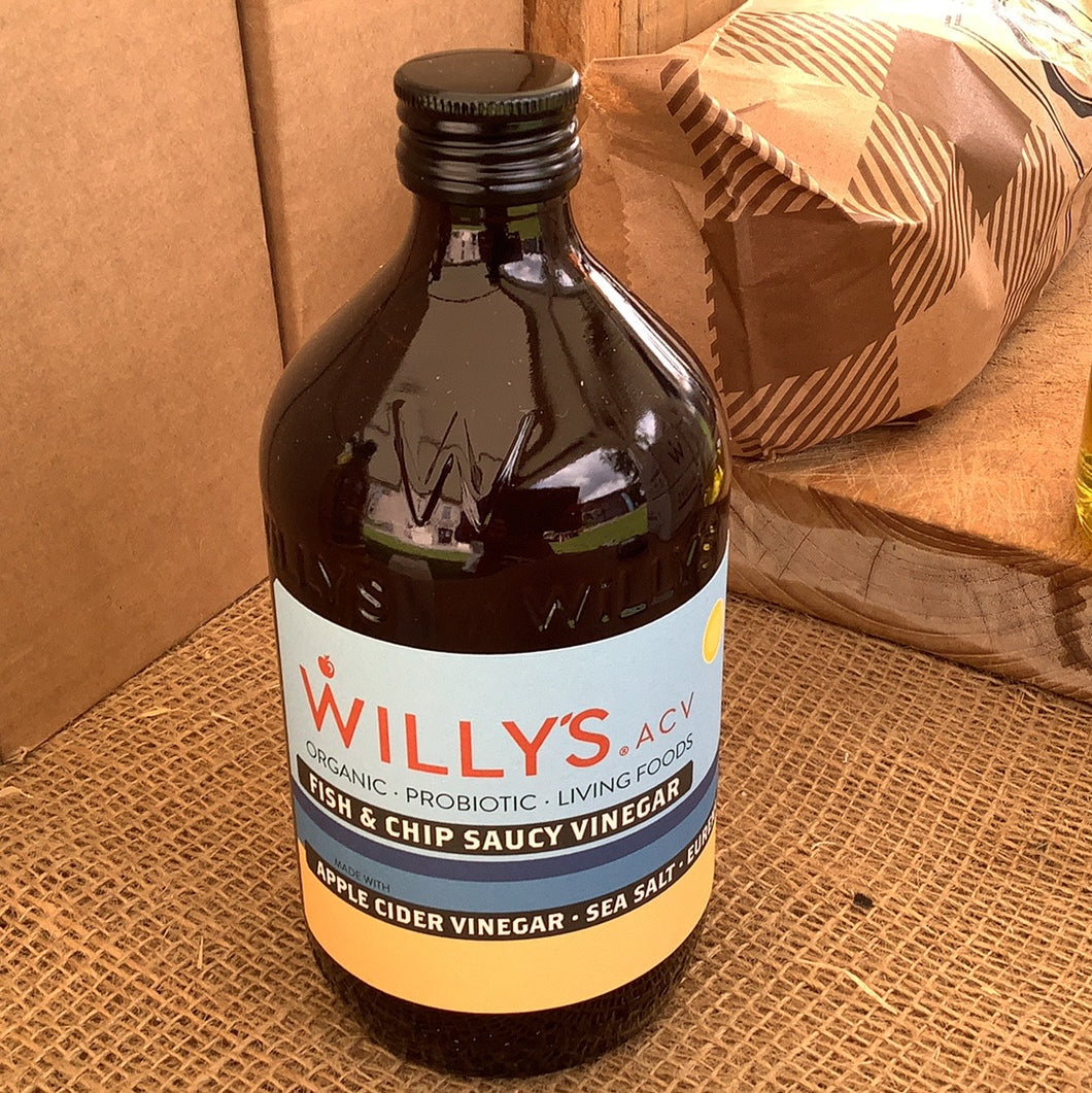 A bottle of Willy's Chip Shop Vinegar with a label that includes the brand name and the words 'Salty Chip Saucy Vinegar'. The bottle is placed on a surface with a cardboard box in the background.